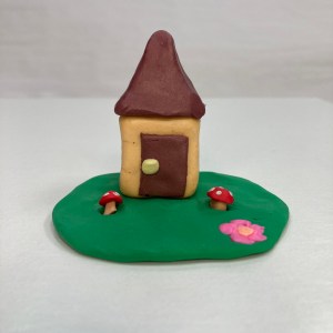 A clay house with one door, atop green landing with a pink flower and mushrooms.