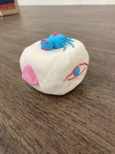 Clay cube decorated with an eye, pink triangle and spider on top.